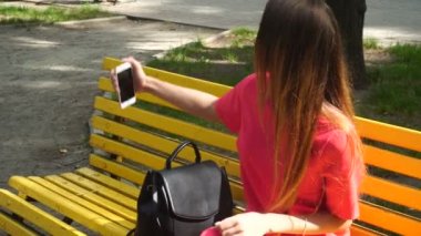 Kırmızı elbiseli genç sarışın kız parkta bir bankta selfie yapar