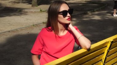 Kırmızı takım elbise ve Güneş gözlükleri çekici genç hanım parkta bir bankta oturur