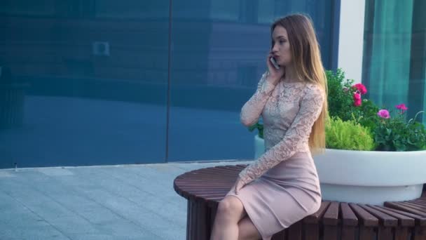 Chica sentada en un banco circular cerca de las flores y habla en un teléfono móvil — Vídeos de Stock