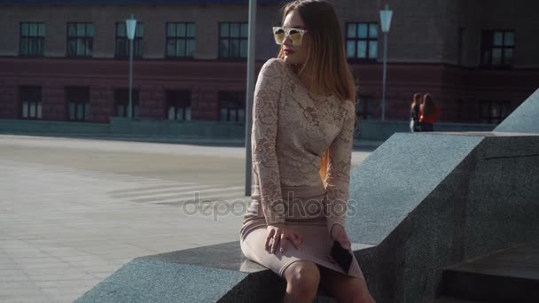 Jovem loira em óculos de sol dourados senta-se na rua — Vídeo de Stock