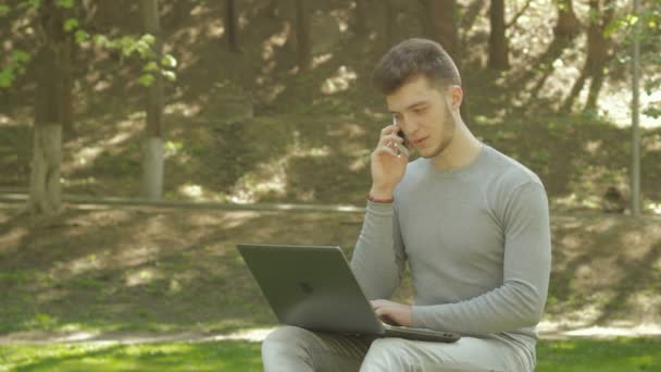 Молодой парень студент в парке разговаривает по телефону и работает с ноутбуком — стоковое видео