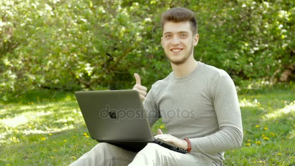 Молодой человек работает с ноутбуком в парке и показывает вверх большие пальцы — стоковое видео