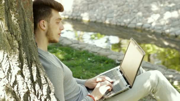 Mann druckt auf Laptop unter Baum im Park — Stockvideo