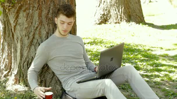 Ragazzo lavora in un parco con un computer portatile e bevitori di caffè — Video Stock