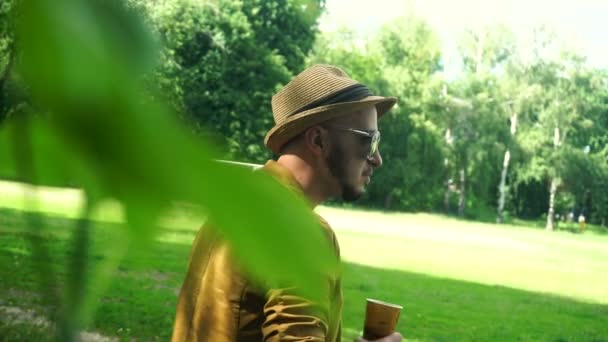 Okouzlující mladý muž v čepici a brýle pije kávu v parku — Stock video