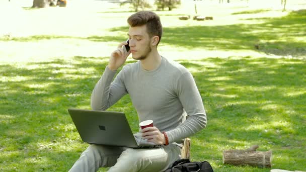 Cara no parque falando ao telefone e bebe café com laptop — Vídeo de Stock