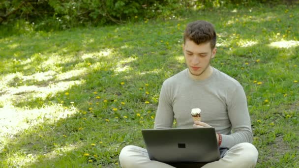 Tipo sentado en la hierba con el ordenador portátil y come un cono de helado — Vídeos de Stock