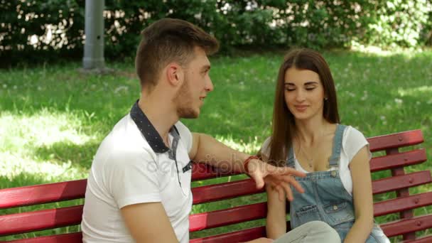 Chico joven coquetea y divierte a la chica en el parque en un banco — Vídeo de stock