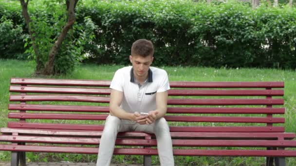 Одинокий парень, сидящий на скамейке с мобильным телефоном в руках — стоковое видео
