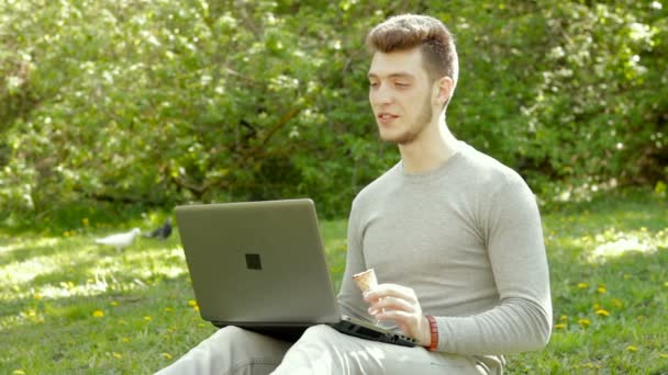 Guapo chico hablando en el video a través de ordenador portátil en el parque y comer helado — Vídeo de stock