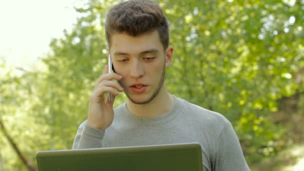 Pretty man waorking with laptop outdoors and talking on mobile phone — Stock Video