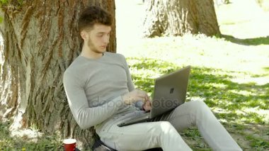 Genç öğrenci sıcak yaz günü ve dizüstü bilgisayar ile çalışma, ağacın altında oturuyor