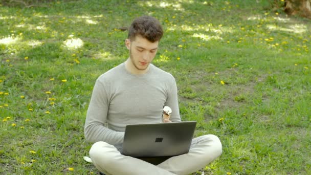 Nadenkende man aan het werk met de laptop op het park en eet en ijs — Stockvideo