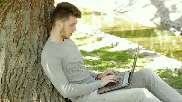 Hombre se sienta a la sombra del árbol trabajando con el ordenador portátil y bebe café — Vídeo de stock