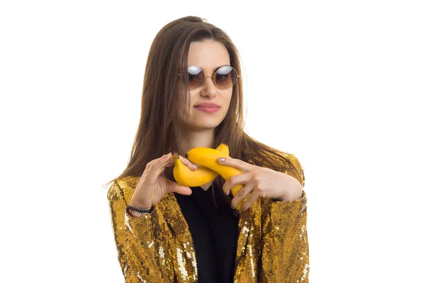 Portret van verbazingwekkende brunette in bril en een gouden jas die in de handen van bananen houdt — Stockfoto