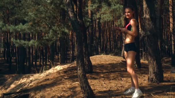 Χαριτωμένο αθλητικό κορίτσι στέκεται κοντά σε ένα δέντρο στο δάσος και κατέχει το τηλέφωνο στο χέρι — Αρχείο Βίντεο