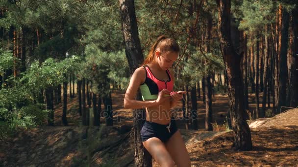 Sportmädchen mit elastischem Bauch steht im Wald und schaut aufs Handy — Stockvideo