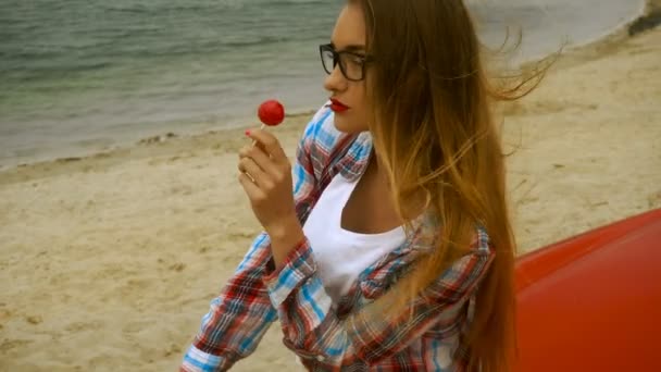 Gorgeous blonde in glasses sits on a red car and licks lollipop — Stock Video