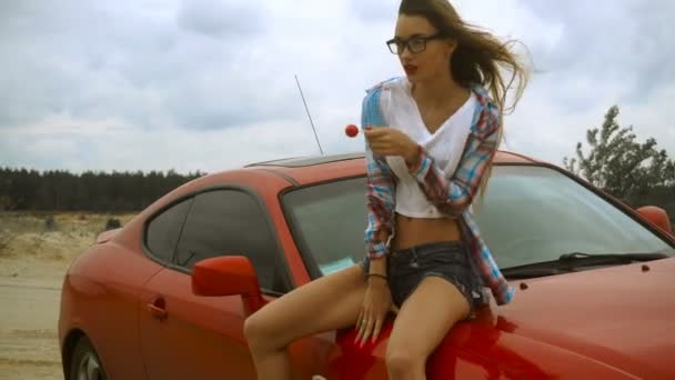 Belleza chica con labios rojos se sienta en un coche y lame una piruleta — Vídeos de Stock
