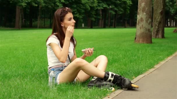 少女はローラーで芝生の上に座っているとインイヤー式ヘッドフォンを挿入します — ストック動画