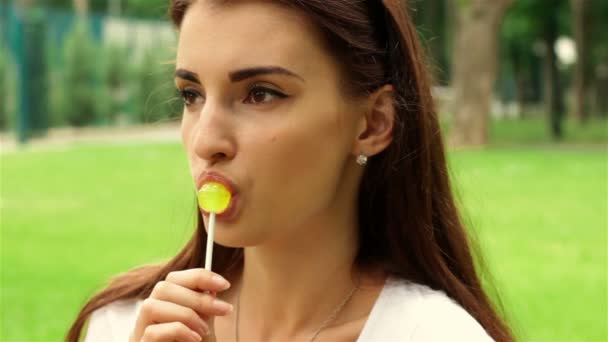 Beautiful girl sits in the Park and licks a lollipop — Stock Video