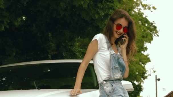 Модна молода брюнетка в окулярах говорить на мобільному телефоні біля машини — стокове відео