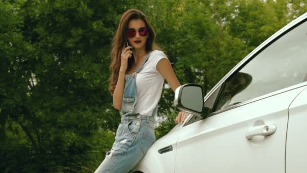 Όμορφη μελαχρινή με φωτεινά γυαλιά στέκεται κοντά στο αυτοκίνητο και λέει τηλέφωνο op — Αρχείο Βίντεο