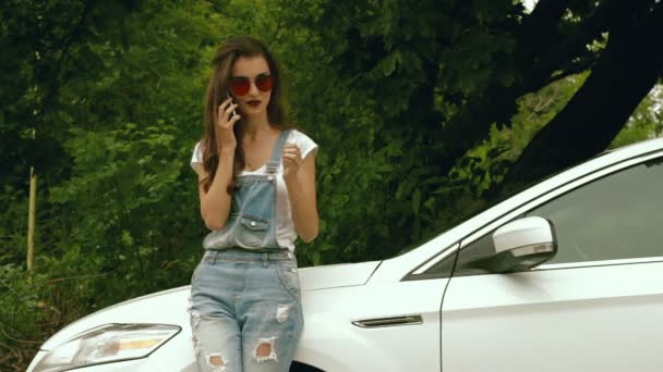 Jolie fille dans des lunettes de soleil lumineuses dit par téléphone près de la voiture — Video