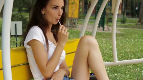 Mladá bruneta sedí na lavičce houpačky a olizuje lízátko — Stock video
