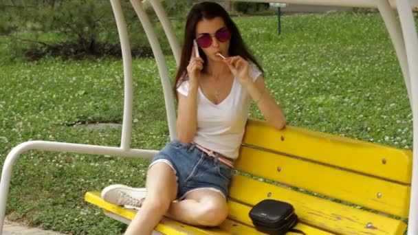 Chica glamorosa en gafas dijo por teléfono en el columpio y lame una piruleta — Vídeos de Stock