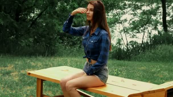 Jovem sexy menina no denim shorts e camisa senta-se no banco — Vídeo de Stock