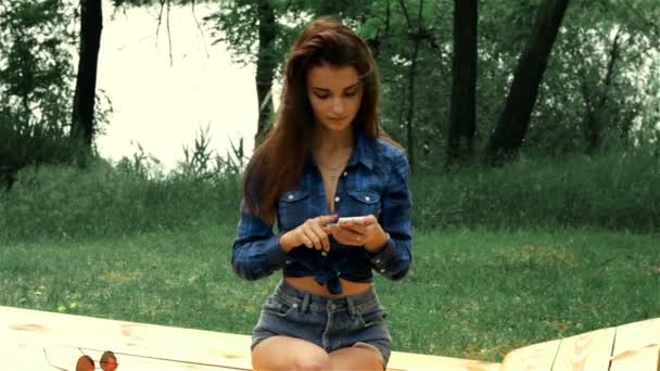 Sottile bruna in camicia seduta su una panchina e guardando al telefono — Video Stock