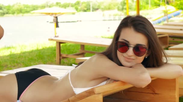 Великолепная юная брюнетка в очках и купальнике лежит в пляжном шезлонге — стоковое видео