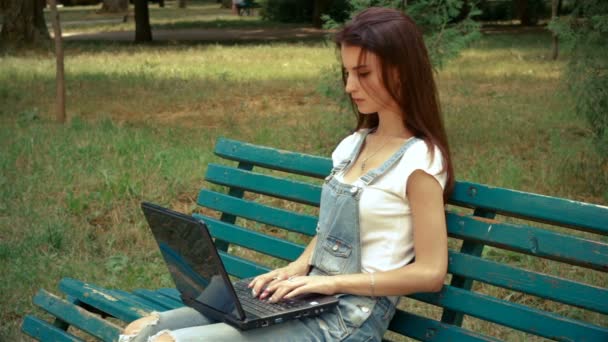 Morena bonito sentado em um banco e impressões em um laptop — Vídeo de Stock