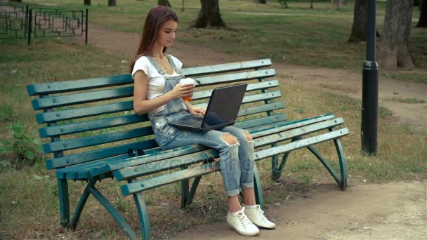 Красива молода дівчина сидить у парку з ноутбуком і п'є каву — стокове відео