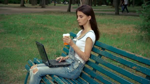 Jovem tem um laptop em suas pernas no parque e beber café — Vídeo de Stock