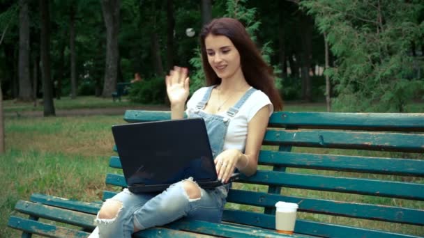 Jeune brune mignonne assise sur un banc avec un ordinateur portable souriant — Video