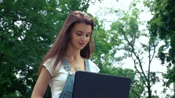 Sorrindo bonito menina olha em um laptop no verão na rua sentado — Vídeo de Stock
