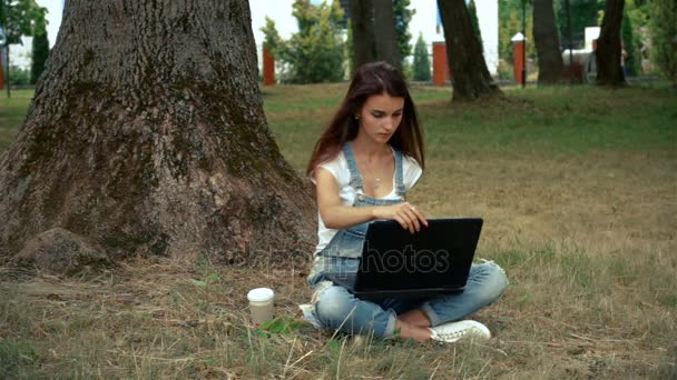 Menina bonito sentado perto de uma árvore no verão e impressões em um laptop — Vídeo de Stock