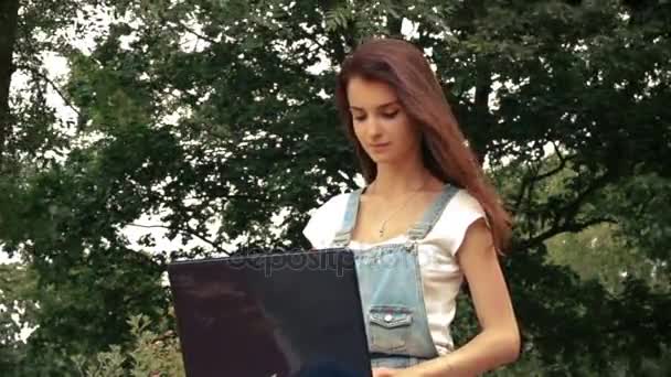 Piękna młoda kobieta siedzi w pobliżu drzew z laptopa i jej włosy lecą — Wideo stockowe