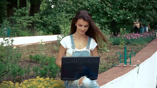 Morena encantadora se senta perto de um canteiro de flores e imprime em um laptop — Vídeo de Stock