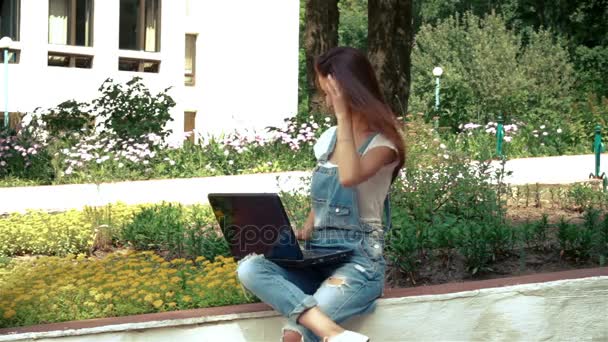 Милая брюнетка в джинсовом комбинезоне сидит на улице и смотрит на ноутбук — стоковое видео