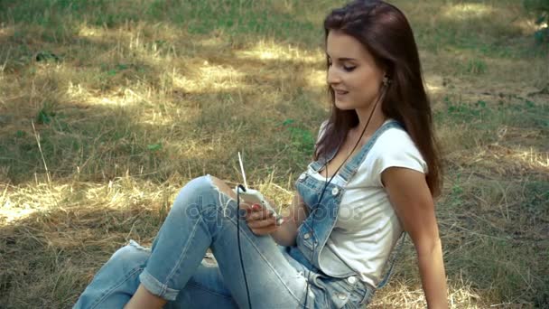 Прекрасная улыбающаяся девушка сидит на траве на улице и слушает музыку. — стоковое видео