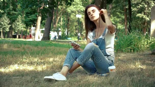 Brunetka, słucha muzyki z słuchawki w telefonie siedzi na trawie — Wideo stockowe