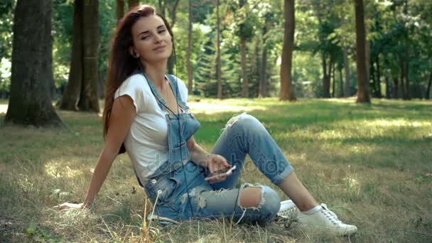 Beautiful girl in jeans suit sitting on the grass outside and listens to music — Stock Video