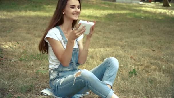 Morena senta-se na grama e segurando uma caixa de presente com um arco — Vídeo de Stock