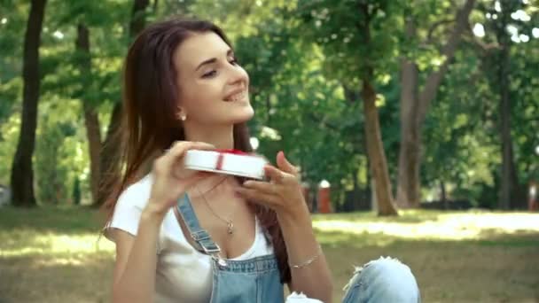 Chica feliz se sienta en el parque en la hierba y la celebración de caja de regalo — Vídeo de stock