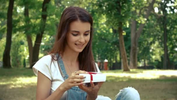 Aranyos lány mosolyog, és úgy néz ki egy kis ajándékot a kezében ül a fűben — Stock videók