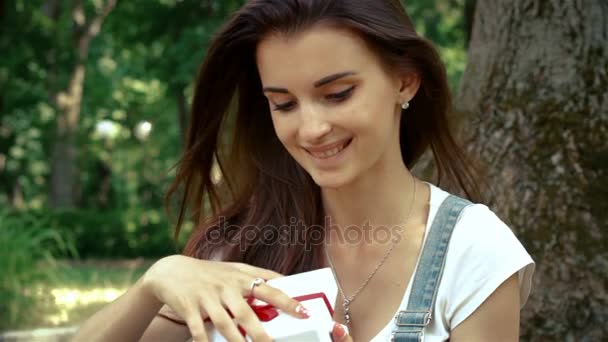 Замечательная девушка сидит на открытом воздухе в парке и открывает подарок — стоковое видео