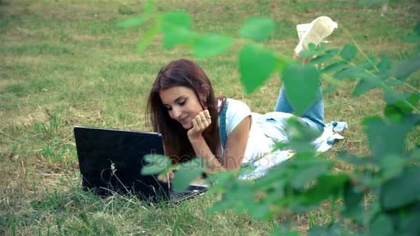 Chica encantadora mira en el ordenador portátil y se encuentra en un parque en la hierba — Vídeos de Stock